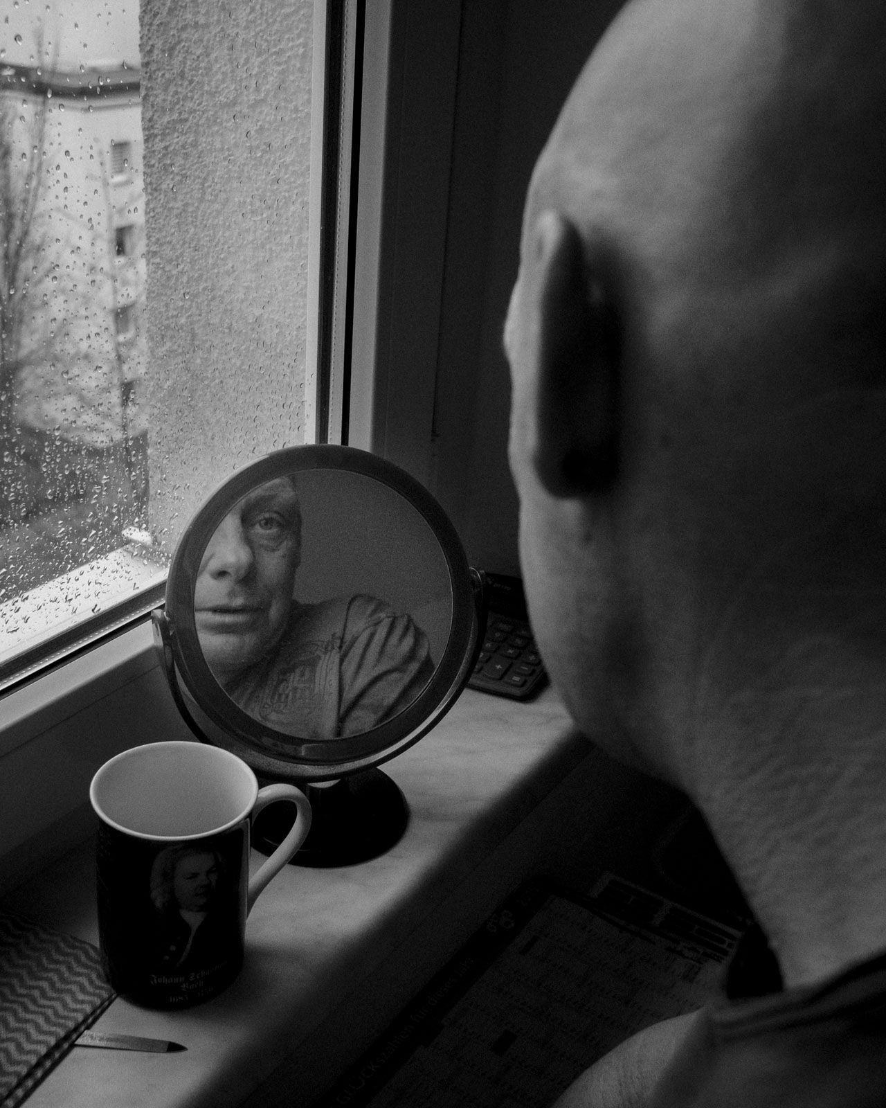 Eike steht am Fensterbrett und betrachtet sich im Spiegel. Er legt Wert auf sein Äusseres, bevorzugt auffällige Kleidung. Die Wohnung ist spärlich eingerichtet: keine Deckenlampen, das Bad von Taschenlampen erleuchtet, Matratze und Waschbecken als einzige Möbel. Fernseher auf dem Boden, ein einzelner Kühlschrank in der Küche. Er sagt, durch seine Nachtschichten brauche er kein Licht. Bad Salzungen, Deutschland, © 2023 Michel Kekulé.