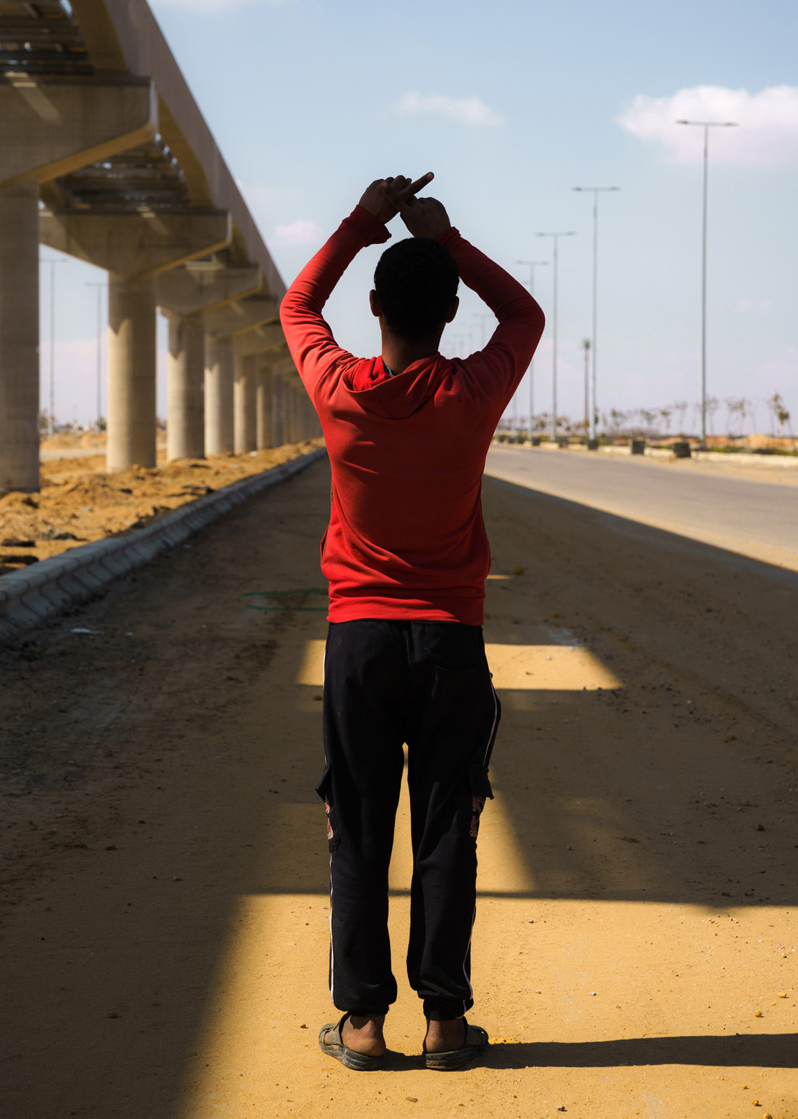 Ein Arbeiter, der täglich die Bewässerungssysteme entlang der mit Rosen gesäumten Straßen der NAC auf Lecks überprüft posiert für die Kamera, Zusammen mit einem Kollegen läuft er durch die Mittagssonne während sie darauf warten von ihrem Teamleiter für die Pause abgeholt zu werden - in einem Pickup Truck mit dutzenden Arbeitern in roten Pullovern hinten drauf, 24.02.2023, New Administrative Capital, Ägypten, © 2023 Louis Roth