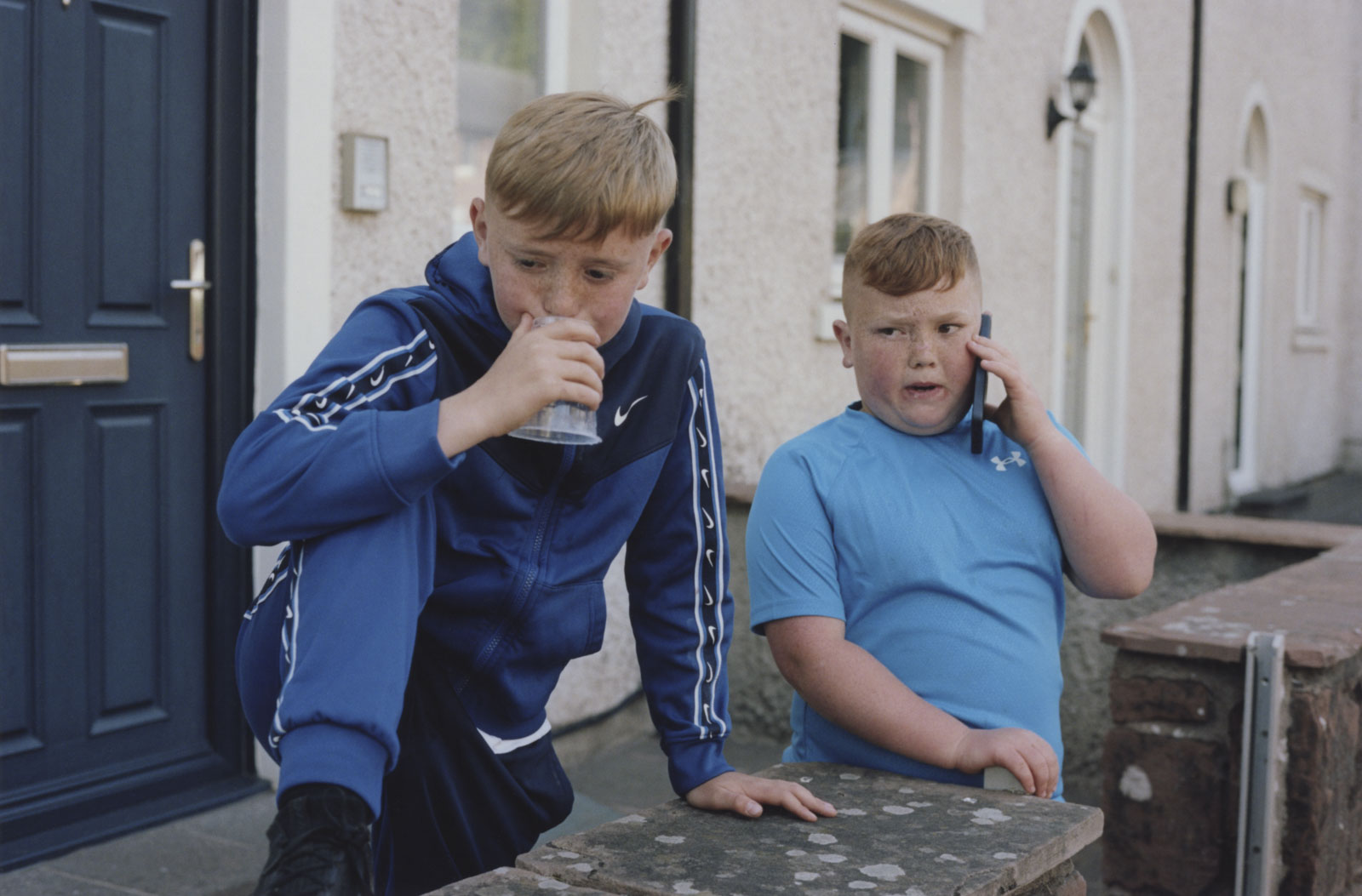 The photo shows two boys. The one on the right side is having a phone call while his friend is holding a cup in front of his mouth. 12.06.2023, Appleby, UK, © 2023 Frederik Rüegger